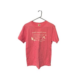 K&ouml;nnte beinhalten: Ein korallenfarbenes T-Shirt mit wei&szlig;em Text und Grafiken. Der Text definiert "CAMPER" und enth&auml;lt eine Definition, einen Stern, einen Baum und eine Zeltgrafik. Das Shirt h&auml;ngt an einem Metallb&uuml;gel.