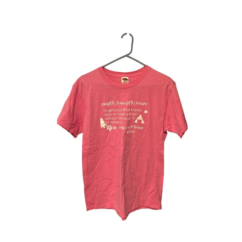 K&ouml;nnte beinhalten: Ein korallenfarbenes T-Shirt mit wei&szlig;em Text und Grafiken. Der Text definiert "CAMPER" und enth&auml;lt eine Definition, einen Stern, einen Baum und eine Zeltgrafik. Das Shirt h&auml;ngt an einem Metallb&uuml;gel.