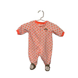 May include: A baby's one-piece pajama set with a white background and orange polka dots. The pajamas have orange trim on the collar, cuffs, and around the feet. A small monkey graphic is on the chest. The feet have a bear face design.