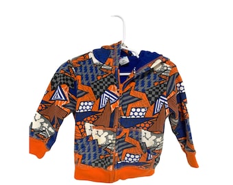 May include: A zip-up hooded sweatshirt with a vibrant geometric pattern in orange, blue, gray, and white. The design includes stars, triangles, and lightning bolts. The cuffs and hem are solid orange, and the interior hood is blue.