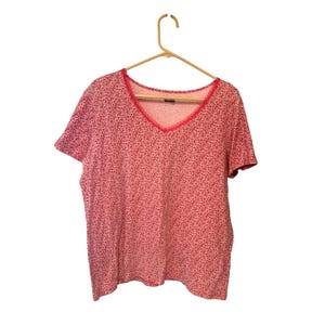 May include: A short-sleeved, V-neck shirt with a floral pattern in shades of pink and red. The neckline is trimmed with a contrasting pink lace. The shirt is hanging on a wooden hanger.