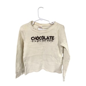 May include: Cream-colored waffle-knit long-sleeve shirt with the text "CHOCOLATE is a girl's best friend" in brown lettering. The shirt is hanging on a white hanger.