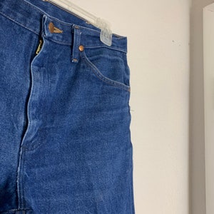 May include: A pair of dark blue denim jeans with a button fly and belt loops. The jeans are hanging on a hanger.