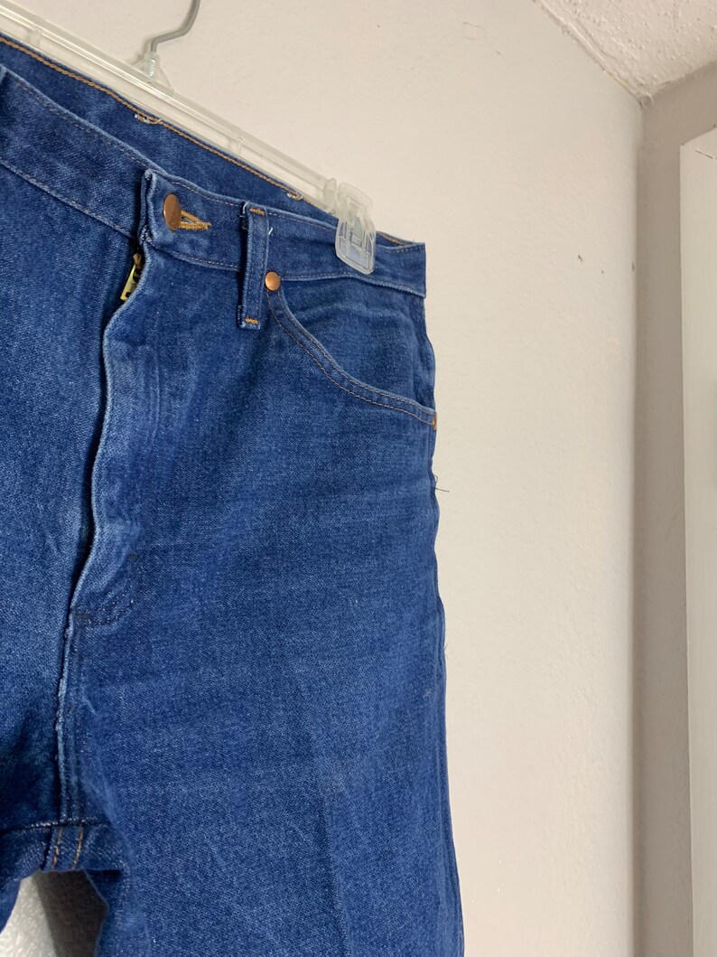May include: A pair of dark blue denim jeans with a button fly and belt loops. The jeans are hanging on a hanger.