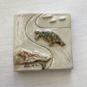 May include: Square ceramic tile with a raised relief design featuring a turtle and a fish swimming in a stylized landscape. The turtle is green and the fish is brown. The tile is off-white with a glossy finish.