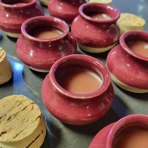 May include: A collection of small, handmade ceramic pots in a deep red hue, filled with a light brown liquid. The pots have a rounded shape with a slightly flared rim and a cream-colored base. Cork stoppers are visible nearby.