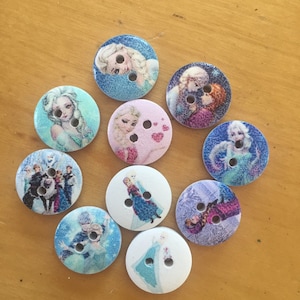 May include: A set of ten round, wooden buttons featuring various images of characters from the animated film Frozen. The buttons are decorated with blue, pink, and white backgrounds and feature images of Elsa, Anna, Olaf, Kristoff, and Sven.