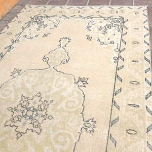 Puede incluir: Una alfombra de color crema con un patrón geométrico en un tono crema más oscuro. La alfombra tiene un borde con un patrón repetitivo de pequeños triángulos y círculos.