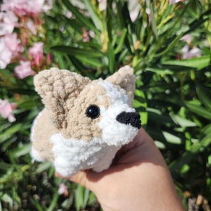 May include: A crocheted stuffed toy dog in the shape of a corgi. The dog is brown and white with black eyes and a black nose. The dog is being held in a hand in front of a bush with pink flowers.