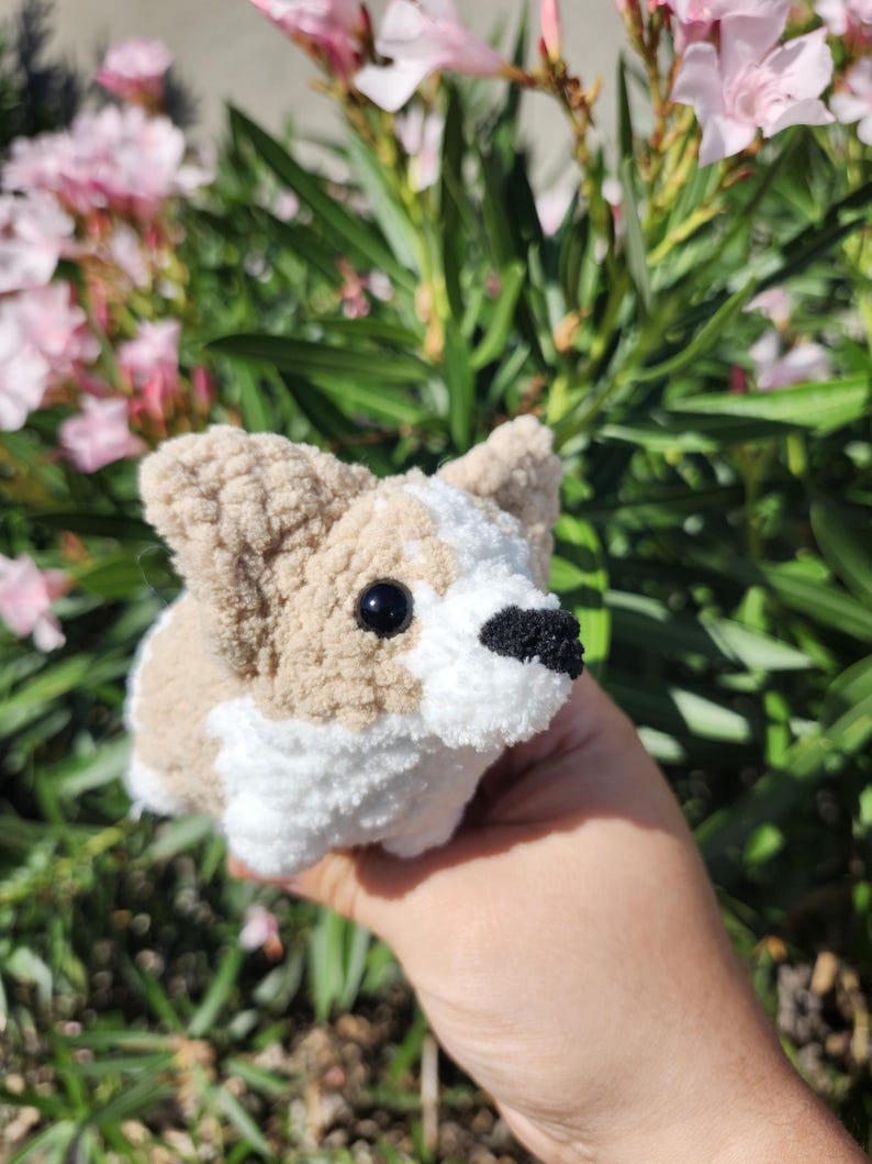 May include: A crocheted stuffed toy dog in the shape of a corgi. The dog is brown and white with black eyes and a black nose. The dog is being held in a hand in front of a bush with pink flowers.