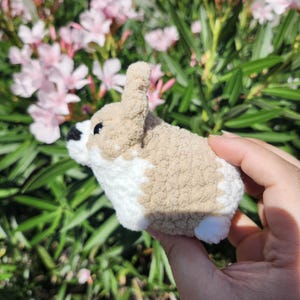 May include: A small, crocheted stuffed animal resembling a dog. The animal is brown and white with a fluffy texture. It is being held in a person's hand.