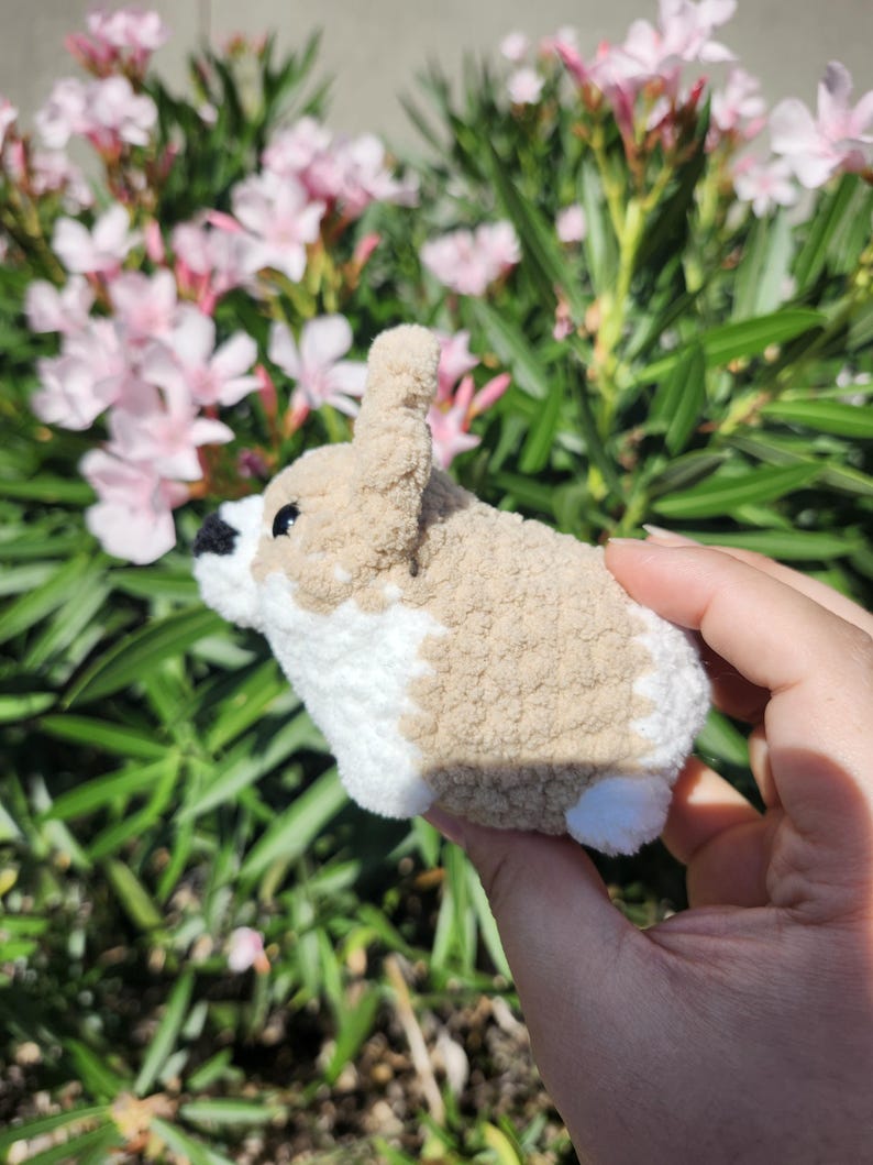 May include: A small, crocheted stuffed animal resembling a dog. The animal is brown and white with a fluffy texture. It is being held in a person's hand.