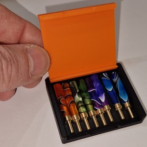 May include: A black box with an orange lid, containing seven colorful pen tips. The pen tips have gold-colored bases and are in shades of orange, brown, green, purple, and blue. The box is being opened by a hand.