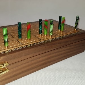 May include: A wooden cribbage board with a walnut finish and a brass inlay. The board has a checkerboard pattern and is closed. There are 12 green, red, and black pegs in the board.