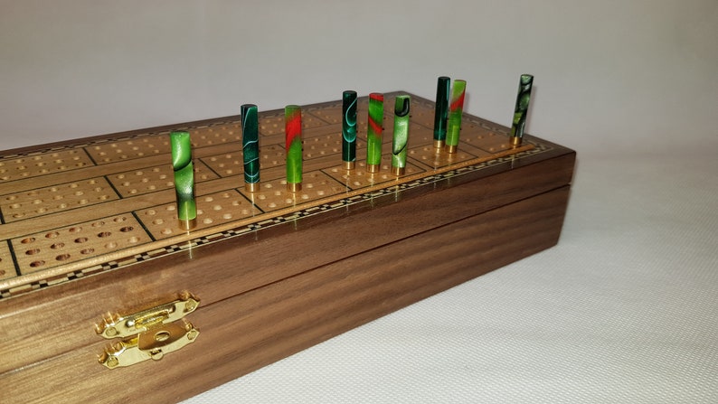 May include: A wooden cribbage board with a walnut finish and a brass inlay. The board has a checkerboard pattern and is closed. There are 12 green, red, and black pegs in the board.