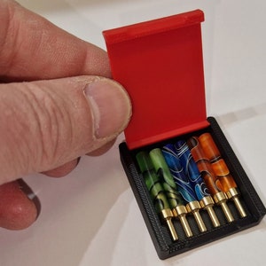 May include: A red, hinged case with a black interior holds six colorful, patterned, and gold-tipped fountain pen nibs.