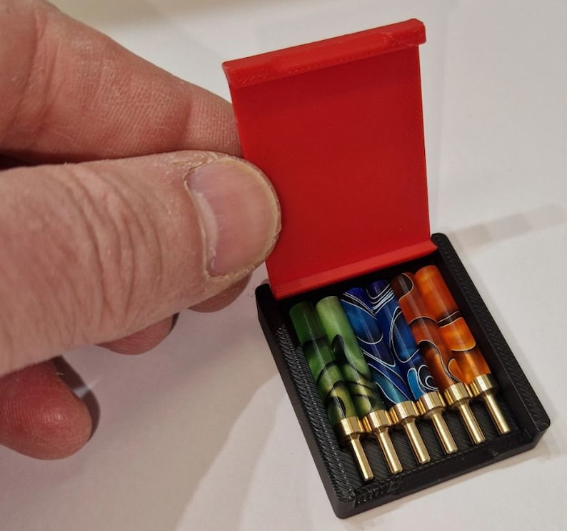 May include: A red, hinged case with a black interior holds six colorful, patterned, and gold-tipped fountain pen nibs.