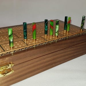May include: A wooden cribbage board with a hinged lid and gold-tone metal latch. The board features a grid of holes for scoring and includes several upright cylindrical pegs made of green and red marbled acrylic with gold-tone metal bases.