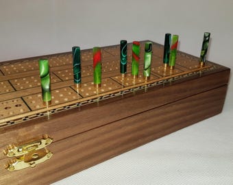 May include: A wooden cribbage board with a hinged lid and gold-tone metal latch. The board features a grid of holes for scoring and includes several upright cylindrical pegs made of green and red marbled acrylic with gold-tone metal bases.