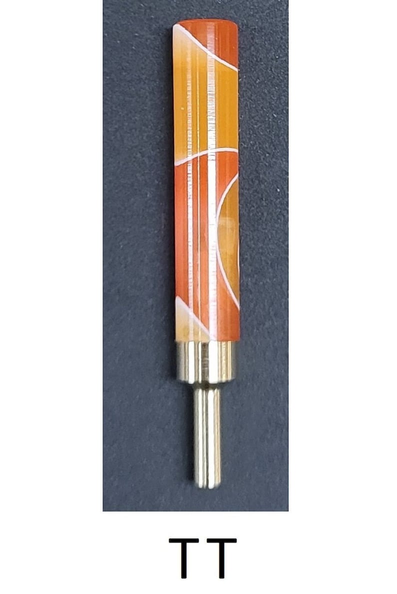 May include: A close-up of a small, orange and white patterned, cylindrical object with a gold metal tip. The object is on a dark gray background. The text "TT" is printed below the object.