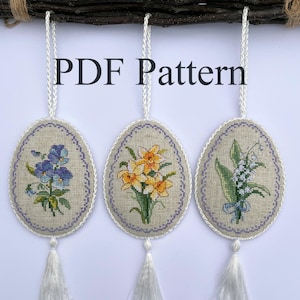 May include: Three oval-shaped cross-stitch ornaments with white tassels hanging from a branch. The ornaments feature embroidered floral designs: purple pansies, yellow daffodils, and white lilies of the valley. The text "PDF Pattern" is printed above the ornaments.