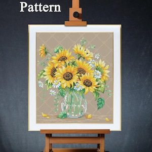 Puede incluir: Patrón de punto de cruz de un ramo de girasoles en un jarrón de cristal transparente. La obra de arte presenta girasoles amarillos y margaritas blancas con hojas verdes. Las palabras "PDF Pattern" están en la esquina superior izquierda. La obra enmarcada se exhibe en un caballete de madera.
