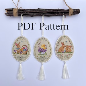 Könnte beinhalten: Drei bestickte Ostereier-Anhänger mit einem Hasen, einem Küken und zwei Hasen. Die Anhänger hängen an einem Zweig und haben weiße Quasten. Der Text "PDF Pattern" ist unter dem Zweig gedruckt.