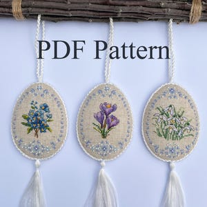 May include: Three oval-shaped cross-stitch ornaments with white tassels hanging from a branch. The ornaments feature floral designs: forget-me-nots, crocuses, and snowdrops.