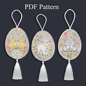 Könnte beinhalten: Drei eiförmige Kreuzstich-Osterornamente mit weißen Quasten und Aufhängeschlaufen. Jedes zeigt ein anderes Design: zwei gelbe Küken, ein Lamm und zwei graue Hasen, alle umgeben von floralen Mustern. Der Text "PDF Pattern" steht oben.