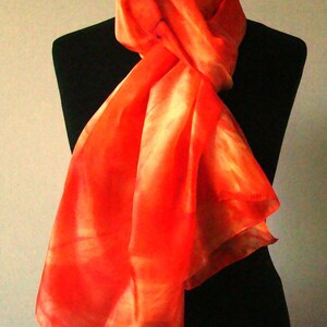 Pode incluir: Um cachecol de seda laranja brilhante com um padrão tie-dye. O cachecol está drapeado sobre um manequim preto.