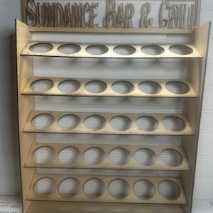 Może przedstawiać: Drewniana półka ekspozycyjna z wieloma okrągłymi otworami na butelki lub słoiki. Na górze półki znajduje się napis "SUNDANCE BAR & GRILL" ozdobną czcionką. Półka wykonana jest z jasnego drewna.