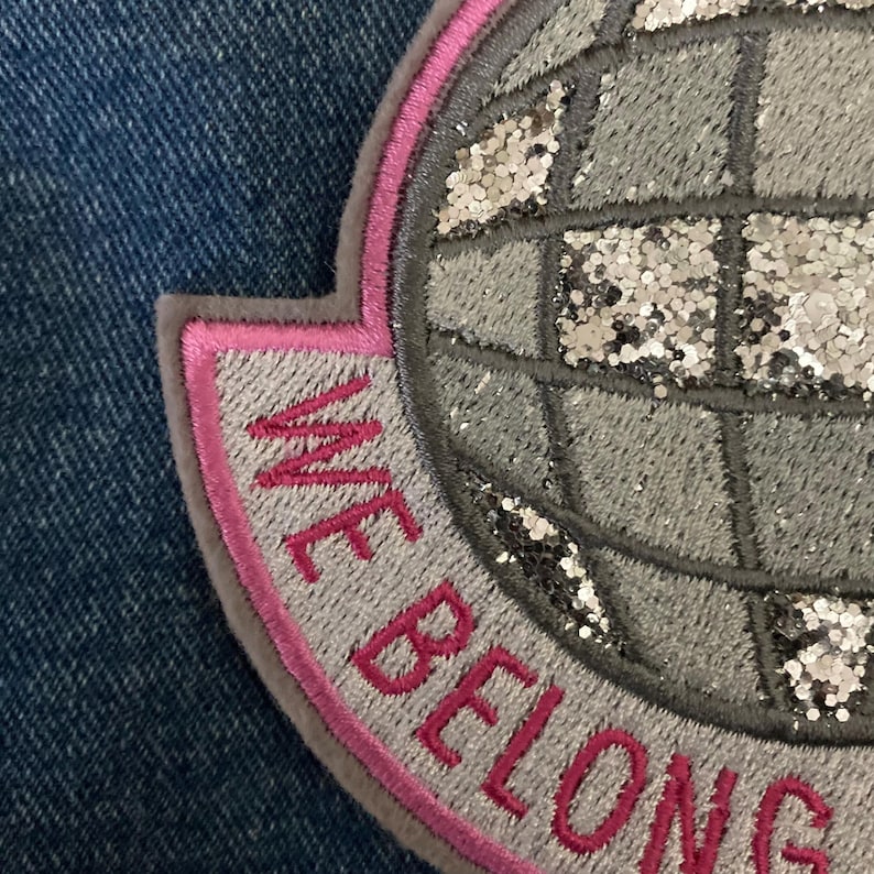 May include: Embroidered patch with the words "WE BELONG" in pink thread. The patch features a gray globe design with silver glitter accents, outlined in pink and gray. The patch is set against a blue denim background.