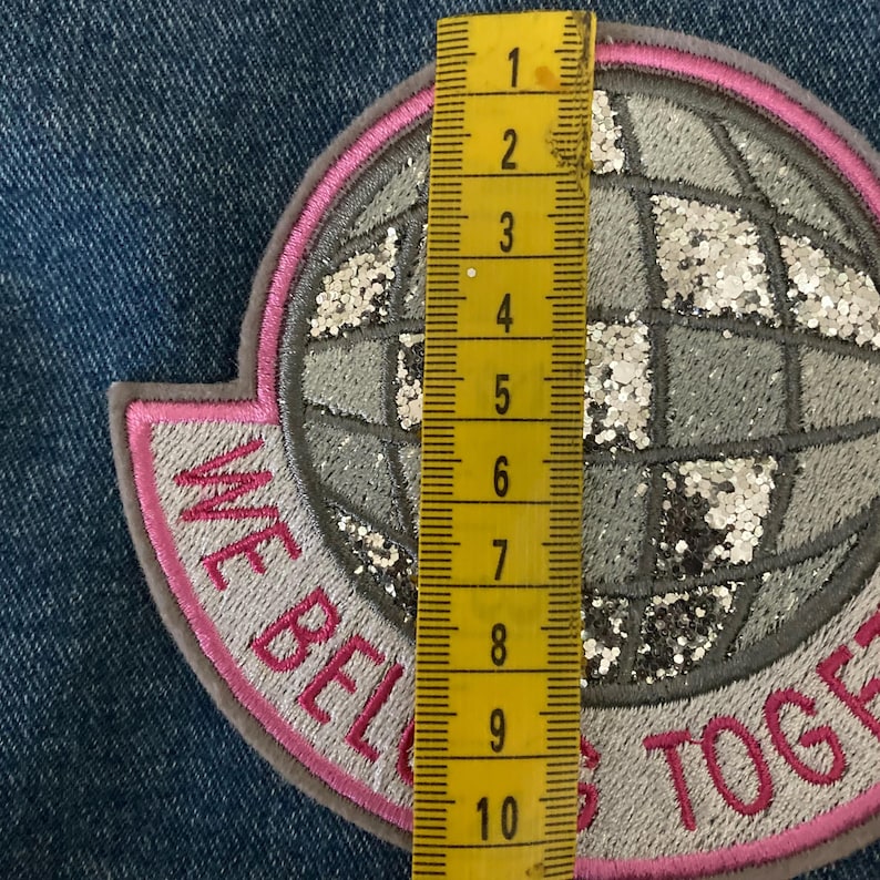 May include: Embroidered patch with a disco ball design, featuring silver glitter and the text "WE BELONG TOGETHER". The patch is approximately 4 inches tall and has a pink and gray color scheme. It is set against a blue denim background.