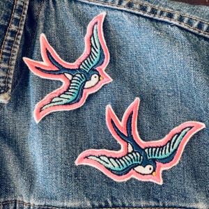 May include: Two pink and blue embroidered swallow bird patches on a denim jacket.