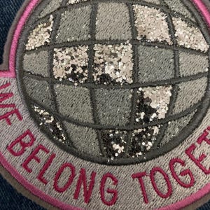 May include: A circular patch with a globe design, featuring gray and silver glitter accents. The patch has a pink border and the words "WE BELONG TOGETHER" embroidered in pink.