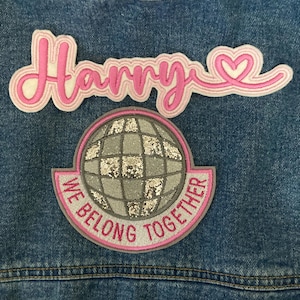 May include: Close-up of a denim jacket with two embroidered patches. The top patch reads "Harry" in pink script with a heart detail. The bottom patch features a silver glitter globe with the text "We Belong Together" in pink.