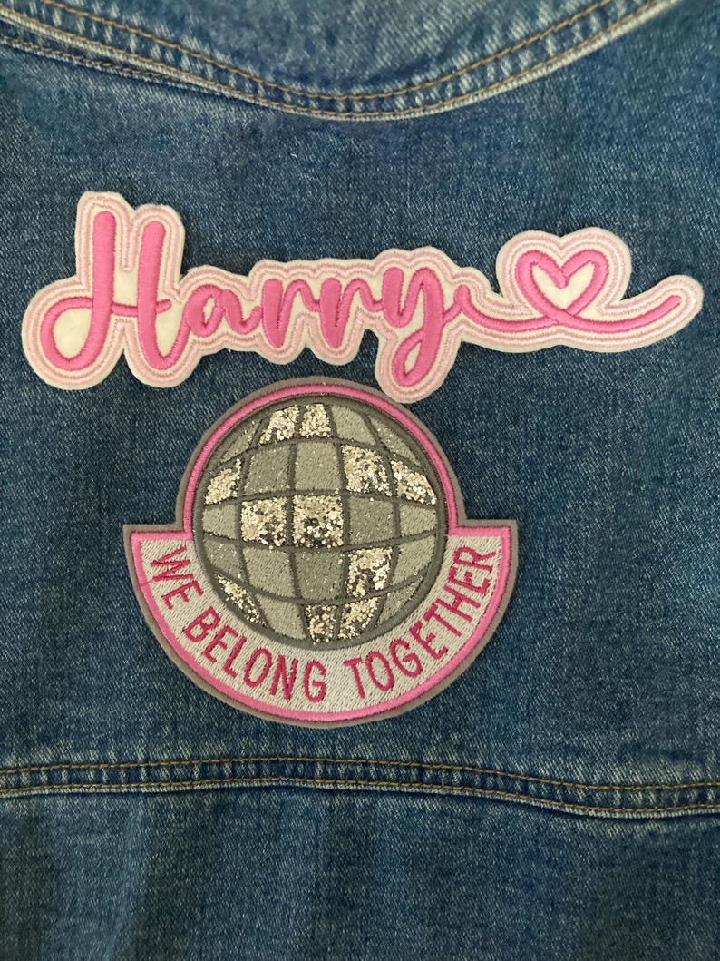 May include: Close-up of a denim jacket with two embroidered patches. The top patch reads "Harry" in pink script with a heart detail. The bottom patch features a silver glitter globe with the text "We Belong Together" in pink.
