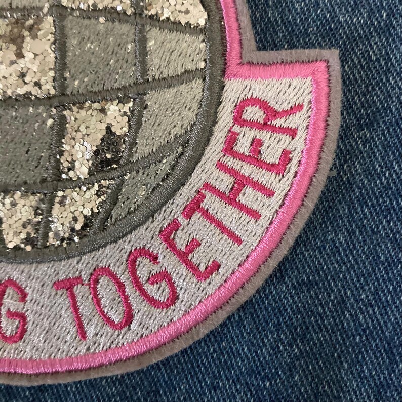 May include: Close-up of an embroidered patch with the words "G TOGETHER" in pink thread. The patch features a globe design with silver glitter accents and a pink border, set against a blue denim background.