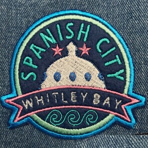 May include: Embroidered patch with a blue, green, and pink colour scheme. It features a white dome with three blue circles, two red stars, and the text "SPANISH CITY WHITLEY BAY" in a navy blue background.