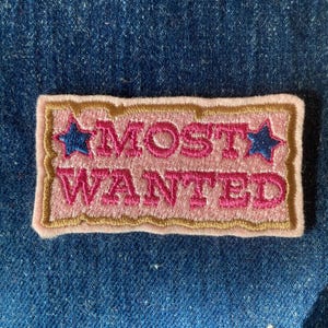 May include: A pink and gold embroidered patch with the text "MOST WANTED" in pink lettering and two blue stars on either side of the text.