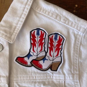 May include: Embroidered patch depicting a pair of cowboy boots. The boots are white with red and blue flame-like designs, brown heels, and a black outline. The patch is sewn onto a white denim jacket.