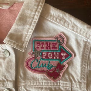 May include: White denim jacket with a pink lining and a "Pink Pony Club" patch. The patch features a turquoise arrow, a pink cowboy boot, and the words "Pink Pony Club" in pink and turquoise.