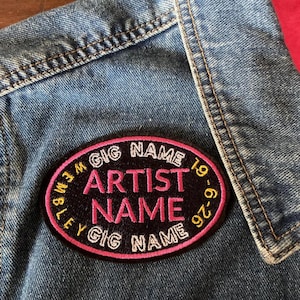 May include: A denim jacket with a pink and black embroidered patch. The patch reads "GIG NAME", "ARTIST NAME", and the date "19-6-26". The oval patch has a pink border and white and gold lettering, sewn onto the blue denim.