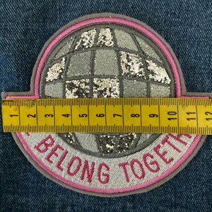 May include: A round patch with a globe design, featuring silver glitter accents and a pink border. The text "BELONG TOGETHER" is embroidered in pink. The patch measures approximately 4.5 inches in diameter.