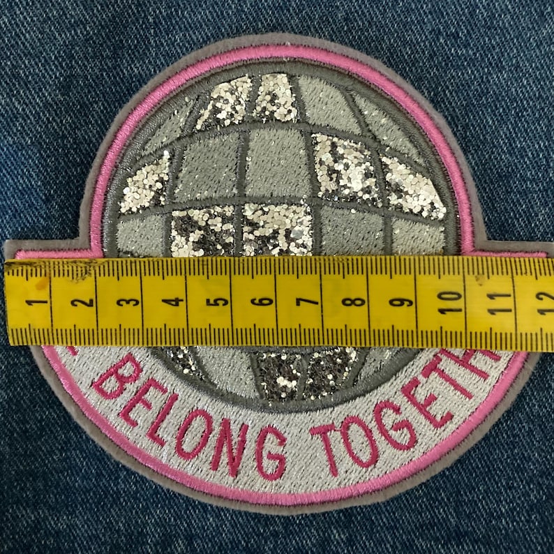 May include: A round patch with a globe design, featuring silver glitter accents and a pink border. The text "BELONG TOGETHER" is embroidered in pink. The patch measures approximately 4.5 inches in diameter.