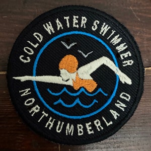 May include: A black and white embroidered patch with a blue circle around a swimmer in the water. The text "COLD WATER SWIMMER NORTHUMBERLAND" is around the circle.