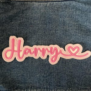 May include: A pink embroidered patch with the name "Harry" in a cursive font. The patch has a white and light pink outline, and a heart design at the end. The patch is on a blue denim background.