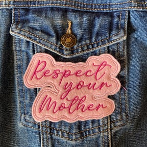 May include: A pink felt patch with the words "Respect your Mother" in cursive lettering is sewn onto a blue denim jacket. The patch has a light pink border and the text is in a darker pink colour. The jacket has a pocket and a button.