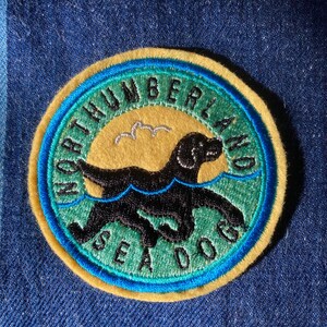 May include: A yellow, blue and green embroidered patch featuring a black dog swimming in the water. The patch reads "Northumberland Sea Dogs".