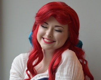 May include: A woman with long red hair wearing a white blouse with puffed sleeves and a blue and white dress. She is smiling and looking down.